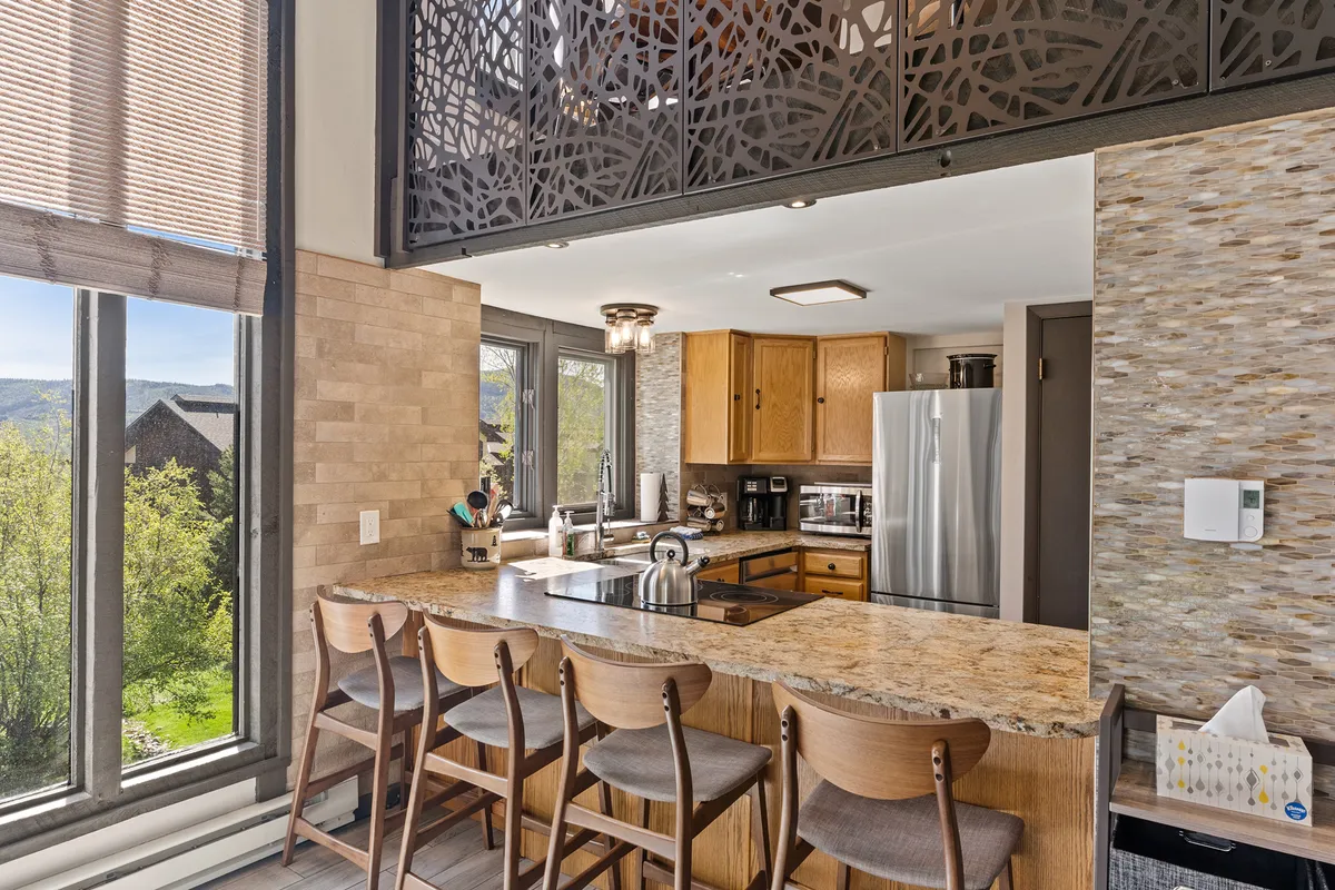 The image showcases a warm and inviting interior with a spacious kitchen island surrounded by stylish bar stools, large windows that fill the space with natural light, and a decorative screen that adds a touch of elegance. The open layout encourages gatherings and socializing, making it ideal for couples or small groups. The overall mood is welcoming, with thoughtfully chosen decor and seating designed for comfort and togetherness.