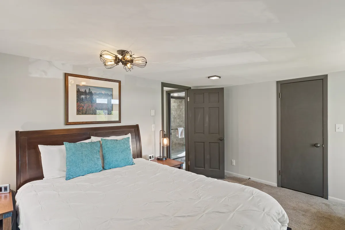 A cozy bedroom with a bed adorned in turquoise pillows and soft linens, with a doorway leading to an adjacent bathroom. Art on the wall and a relaxing ambiance make the space feel like a restful sanctuary.