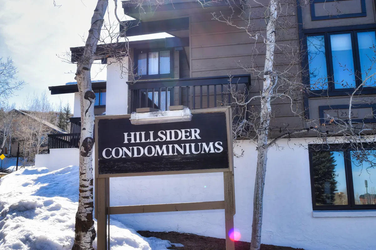 The image captures the Hillsider Condominiums sign flanked by snowbanks, with the building’s distinct architecture visible behind. Snow blankets the landscape, indicating close proximity to winter slopes, and the atmosphere is welcoming and cozy.