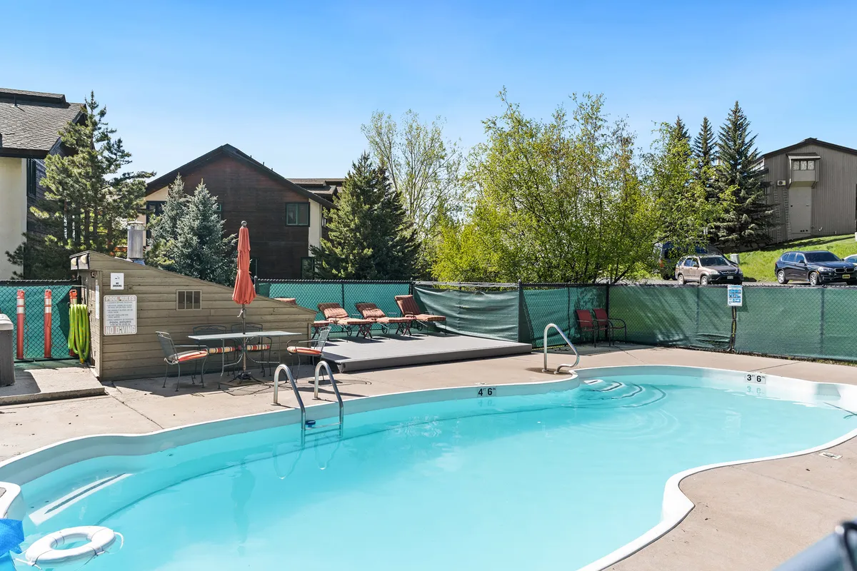 This image features a sparkling community swimming pool surrounded by lounge chairs and a table shaded by an umbrella. Mature trees create a lush, inviting backdrop for summer relaxation. The setup invites guests to imagine themselves swimming, sunbathing, or enjoying a meal by the pool.