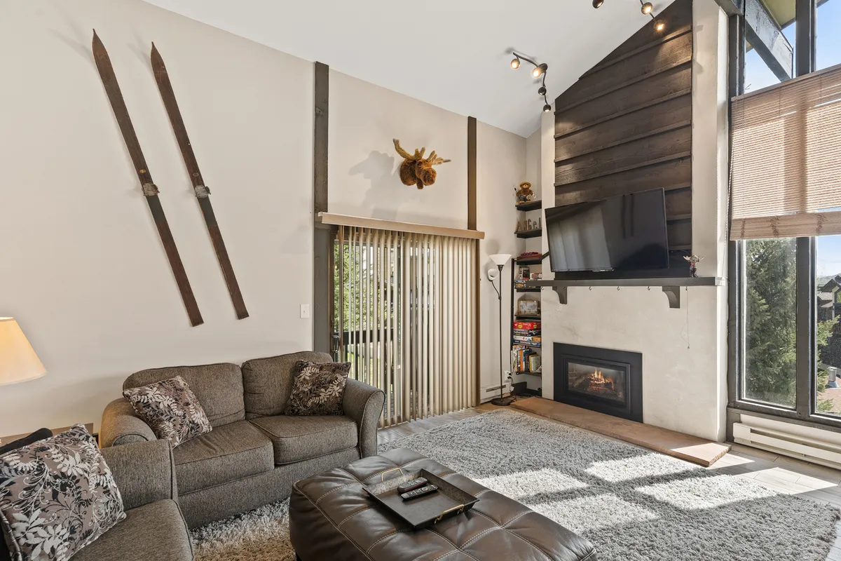 The image features a welcoming living room with a plush sofa set, a gas fireplace as the central focal point, and mountain-themed decor including wooden skis. The layout feels warm and homey, with ample space for gathering and relaxing after outdoor adventures.